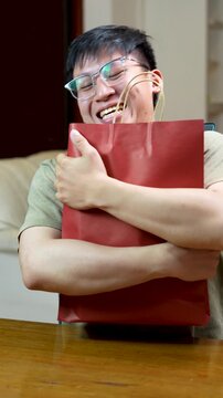 Vertical shot of an ecstatic happy young Asian man hugging a red paper shopping bag tightly and laughing with joy indoors