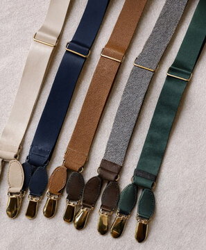 Leather and fabric suspender straps in cream, navy, brown, gray, and green arranged on a light textured surface. Classic accessories for formal styling.