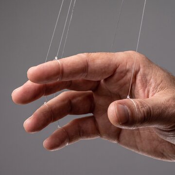 Close up of a human hand with fingers tied to clear strings like a marionette puppet illustrating psychological manipulation and control
