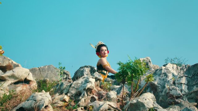 A cinematic low angle shot of a woman in Javanese traditional attire sitting perched on the top of a jagged limestone rock formation against a clear blue sky.