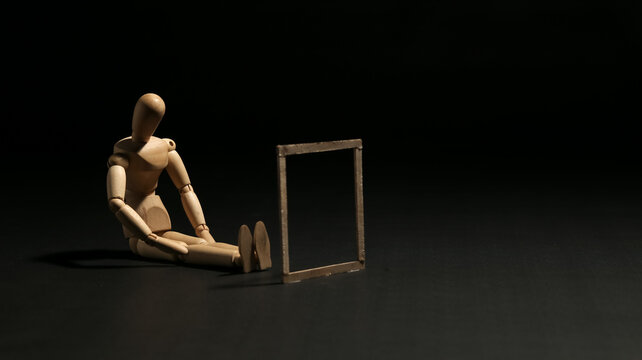 Wooden mannequin sitting on the floor looking at an empty frame. Concept of self reflection, loneliness, missing piece, perspective, future vision or memory.