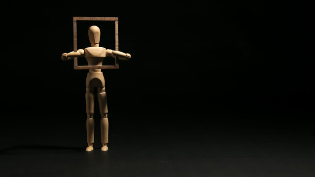 Wooden mannequin holding a wooden square frame around its head and chest. Concept of framing, perspective, identity, boundaries, limitation or thinking inside the box.