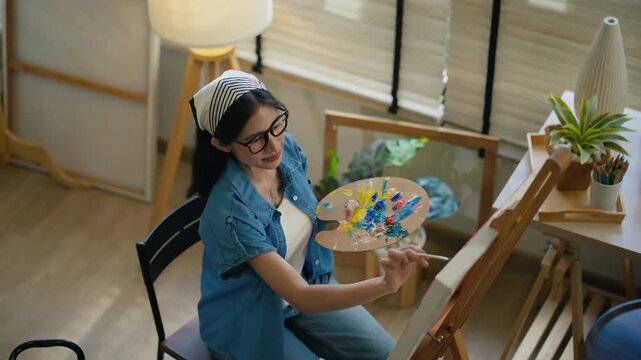 Gen Z Asian woman freelance artist painting with paintbrush on canvas easel, representing creative professional women designer, online independent creator and art studio hobby lifestyle