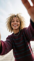 Fototapeta premium Happy young woman with curly blonde hair takes selfie on wooden pier. Smiling girl wears striped sweater, blue necklace. Joyful person enjoys summer vacation near ocean shore. Beautiful female