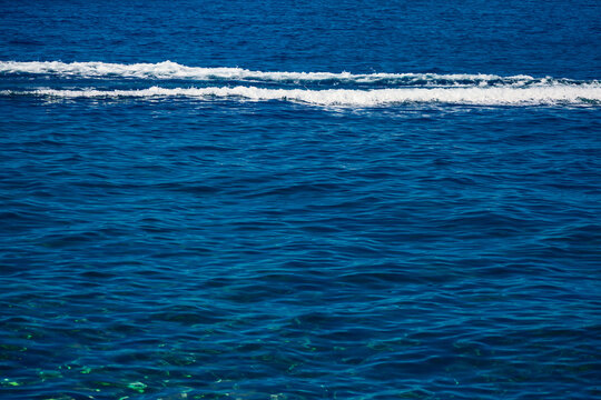 Boat leaves a long, foamy wake across deep ocean. Ripples spread outward from moving vessel, creating simple, textured seascape. Gentle ocean waves form thin white lines across calm, rippling surface