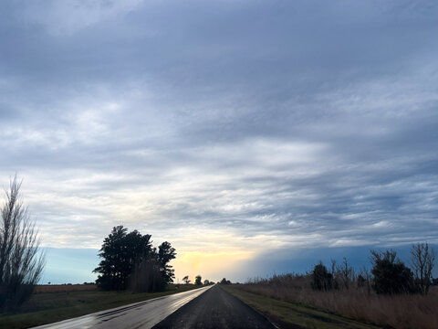 Storm clouds forming over a dry agricultural field, with a dramatic dark sky and light rain falling in the distance, illuminated by the bright golden light of the setting sun on the horizon.