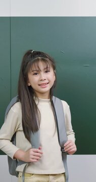 Cute Asian schoolgirl with backpack smiling happily in classroom.