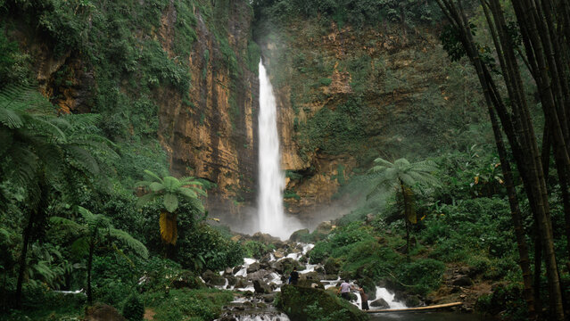 Center composition of tall waterfall cascading down steep cliff surrounded by lush green tropical forest. Stream flowing over rocks in foreground with tall trees framing both sides.