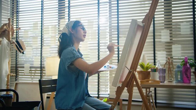 Gen Z Asian woman freelance artist painting with paintbrush on canvas easel, representing creative professional women designer, online independent creator and art studio hobby lifestyle