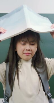 Playful Asian schoolgirl hiding face behind a notebook and smiling at camera