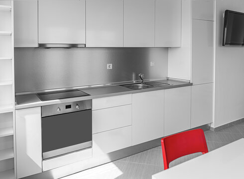 A modern minimalist white kitchen features sleek cabinets, stainless steel appliances, and a single red chair for a splash of color.