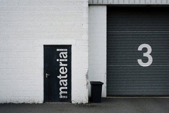Industrial warehouse exterior with white brick wall, black metal door. Large door features white material text. Grey rolling shutter garage gate shows number 3. Black trash bin sits on concrete