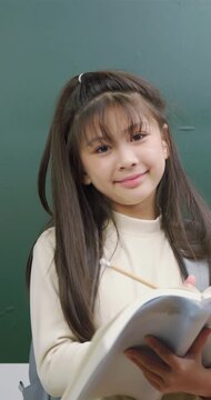 A cute young girl holding an open book and a pencil in front of a green chalkboard. She has a bright idea, points her pencil up, and smiles happily.