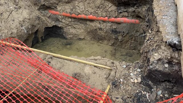 Excavation pit dug in asphalt road filled with murky water next to orange safety netting at construction site: revealed underground laying of sewer pipe. Muddy trench shows layers of soil, earth