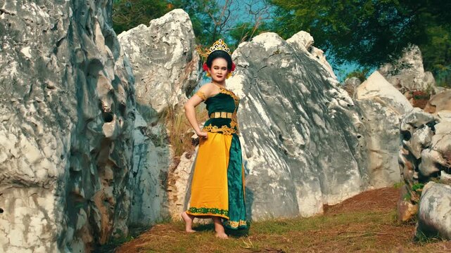 A full-body shot of a confident Indonesian woman wearing a traditional green and yellow dance costume, standing barefoot and posing with hands on hips.