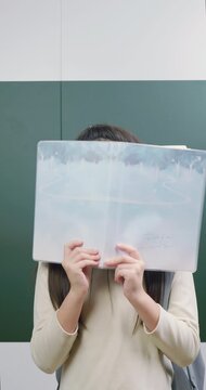 Playful Asian schoolgirl hiding face behind a notebook and smiling at camera