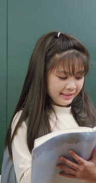 A cute young girl holding an open book and a pencil in front of a green chalkboard. She has a bright idea, points her pencil up, and smiles happily.