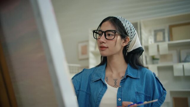 Gen Z Asian woman freelance artist painting with paintbrush on canvas easel, representing creative professional women designer, online independent creator and art studio hobby lifestyle
