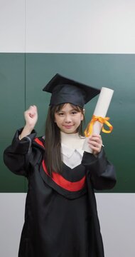 Cute Asian graduate girl holding diploma scroll celebrating success with raised fist gesture.