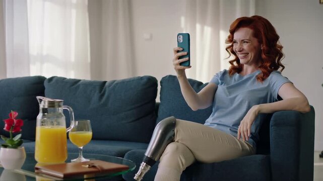 Woman with prosthetic leg sitting on blue sofa using smartphone for video call in living room