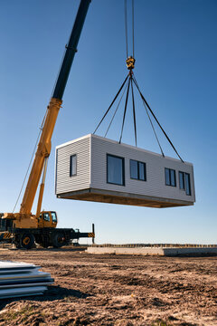 A yellow crane lifts a prefabricated house module with gray siding. It is suspended over a concrete foundation on a dirt construction site