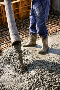 A person stands on a construction site pouring concrete from a hose onto a metal framework. The sun shines down making the scene bright and busy