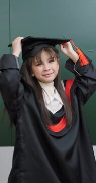 A young girl wears a graduation gown and cap. She smiles happily in front of a chalkboard.