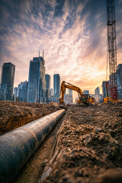 Significant excavation is underway at a construction site in the city with a long dark pipeline laid in a trench, while a yellow excavator works