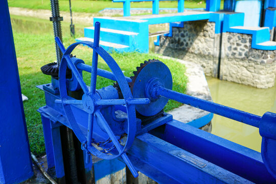 Blue mechanical water sluice gate mechanism with bevel gears and threaded rod at an outdoor irrigation canal system, controlling river water flow for agriculture and flood control management.