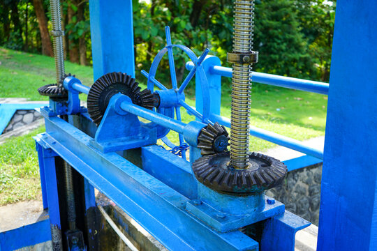 Blue mechanical water sluice gate mechanism with bevel gears and threaded rod at an outdoor irrigation canal system, controlling river water flow for agriculture and flood control management.
