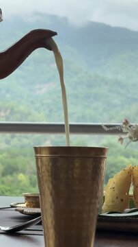 Pouring fresh lassi into traditional brass glass with scenic mountain background vertical video
