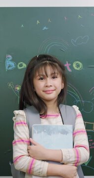 Cute Asian schoolgirl with backpack smiling happily in classroom.