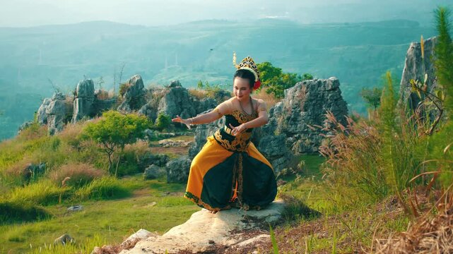 A scenic wide shot of a traditional dancer in a low stance on a rock formation, with a backdrop of green hills and a misty horizon.