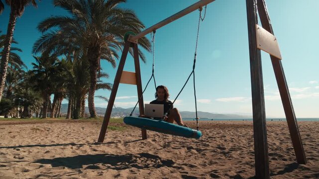 Young man with long hair working on a laptop while sitting on a blue swing on a sandy beach looking out at the ocean, swing set, swing chains, laptop lifestyle, digital nomad
