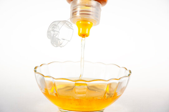 Golden Natural Honey Dripping from Squeeze Bottle into Clear Glass Bowl on Pure White Background