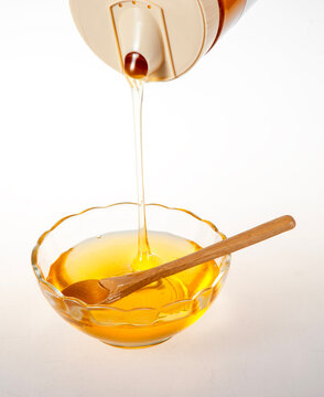 Golden Raw Honey Dripping from Squeeze Bottle into Glass Bowl with Wooden Spoon on White Background