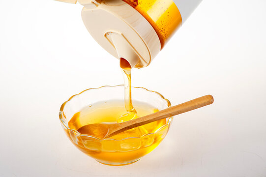 Golden Raw Honey Dripping from Squeeze Bottle into Glass Bowl with Wooden Spoon on White Background