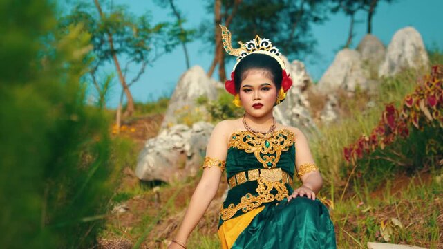 A beautiful Indonesian woman wearing a traditional green velvet Kebaya sitting gracefully on the ground amidst limestone rocks and trees.