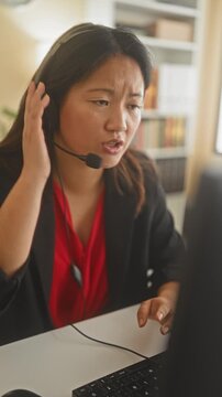 Woman agent holds headset to ear and types at keyboard in an office building, frowning while speaking into mic; customer support frustration.