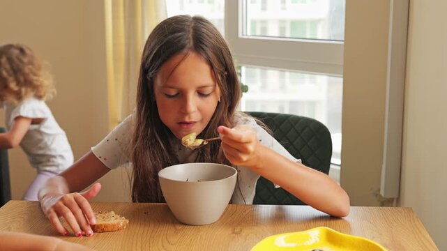 Young girl eating a slice of bread with her breakfast at a wooden dining table in a bright modern kitchen at home.