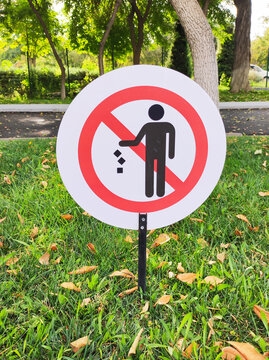 Round white prohibition sign with red border and crossed out person dropping trash icon on black pole. Standing in green grass with fallen leaves in city park. Ecology and public rules concept.