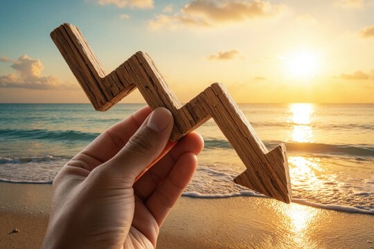 Hand holds wooden downward arrow graph at sunset beach. Economic crisis concept with declining market trend, financial loss. Business recession stock crash shown by falling chart against ocean