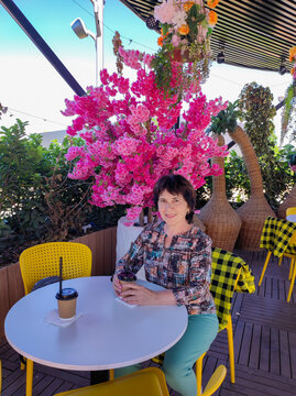 Smiling mature woman sitting at round white table in outdoor summer cafe holding coffee cup. Bright pink flowering tree and yellow chairs in background.