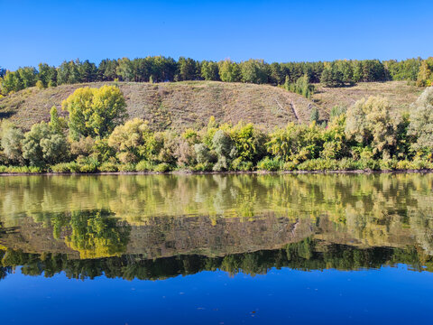 Calm river reflecting a grassy hill and mixed forest under a clear blue sky. Early autumn landscape with yellowing trees, applied for travel, tourism, and ecology design backgrounds.