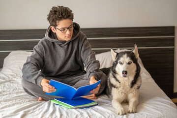 Young student writing homework while husky sits close, dog sitting exactly where notebook lies, moment impossible to repeat on second day. Wide shot. Concept of homework done together with own pet © iwavephoto