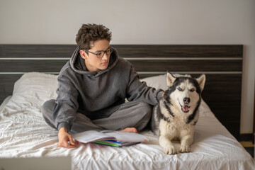 Young student writing homework while husky sits close, dog sitting exactly where notebook lies, friendship without any condition. Wide shot. Concept of homework done together with own pet © iwavephoto