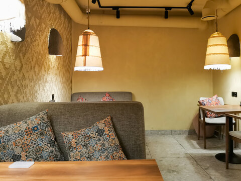Empty eastern cafe interior with gray booth seating and colorful oriental patterned pillows. Wooden tables and hanging pendant lamps illuminate the cozy dining space for hospitality and food service.