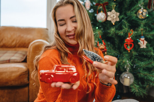 Woman holds red toy car and decoration near Christmas tree in cozy