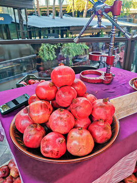 Pile of fresh red pomegranates in clay bowl on a purple tablecloth at a market stall. A manual juicer press stands nearby. Fresh produce, healthy food, and street bazaar concept.