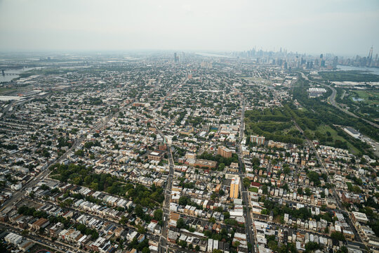 An aerial view shows a dense residential neighborhood with streets and houses stretching across the city. The Manhattan skyline rises in the distance creating a dramatic cityscape panorama.
